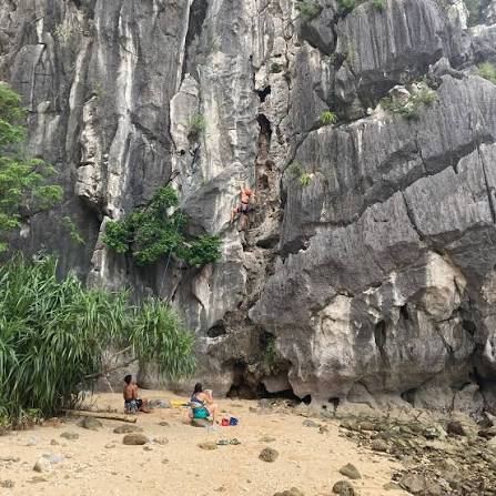 A climber on a limestone wall with participants on the sandy beach below